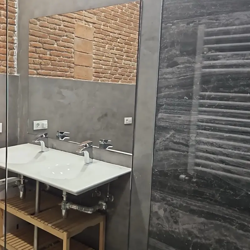 Baño de microcemento con pared de ladrillo visto, espejo a medida y doble lavabo sobre estructura de acero