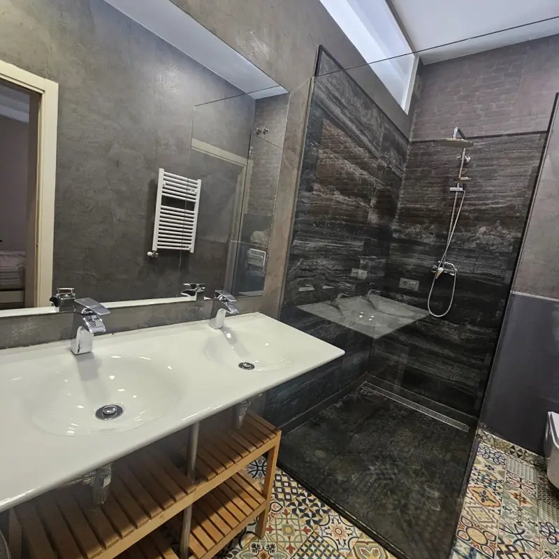 Baño de microcemento gris con ducha de obra de porcelánico efecto mármol oscuro, doble lavabo y suelo hidráulico