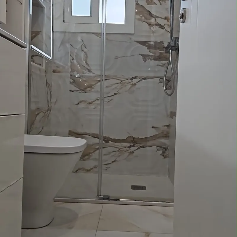 Ducha de obra con plato a ras de suelo, mampara corredera de cristal y porcelánico efecto mármol, con inodoro al lado y ventana al fondo